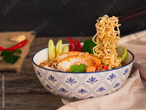 Use chopsticks to pick up ramen instant noodle soup with shrimp in white bowl on wooden table background. Asia Food, Tom Yum Kung