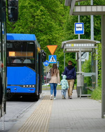 Family with a child walking towards a modern bus stop shelter on a city street as a blue public bus arrives, illustrating urban daily life and transportation