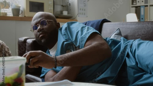 Sleepy African American male clinician in medical scrubs napping on couch in break room, taking mobile gadget from table, getting up and hurrying to sick patient