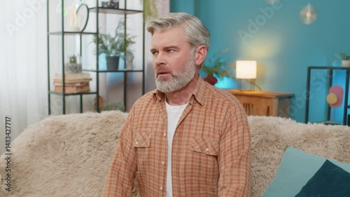 Caucasian mature man sitting on sofa at home, holding smartphone and showing signs of stress. Middle-aged guy receives bad news message, feeling despair and exhaustion, looking tired overwhelmed