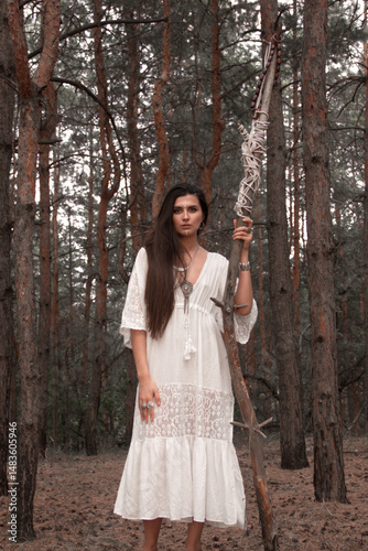 young brunette girl in white boho style dress is stands casual with long high wooden witch staff in hand and looks straight on a pine forest background. rustic esoteric. lifestyle concept, free space