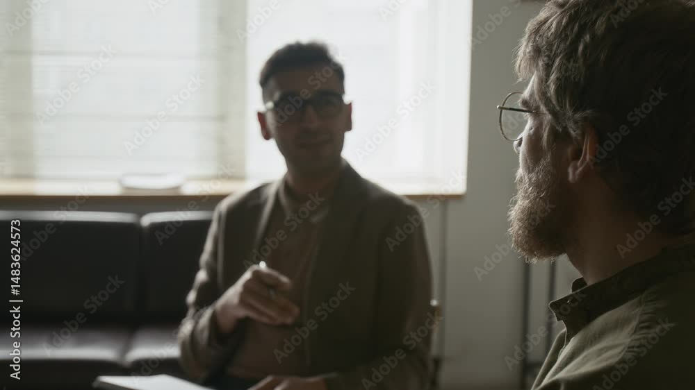 Indian male therapist speaking and leading discussion while one of group therapy participants listening attentively in focused foreground, going through healing in calm atmosphere in softly lit room
