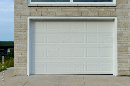 A large white metal car garage door on a grey brick house. The solid single steel door has square patterns or panels. The modern luxury automotive building has a grey concrete driveway entrance. 