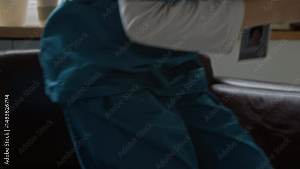 Young sleeping female clinician in medical scrubs lying on sofa, then taking calling mobile gadget from table, getting up and moving towards door of break room