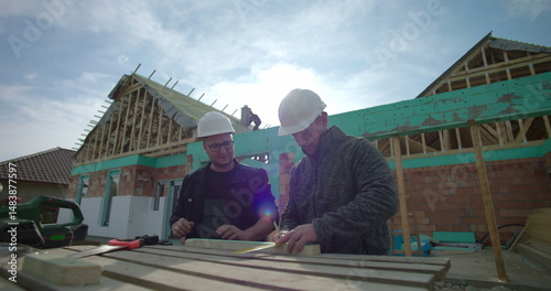 Close-up of two builders marking wood, teamwork and accuracy, house construction, residential project in progress, hard hats, worktable with tools and materials