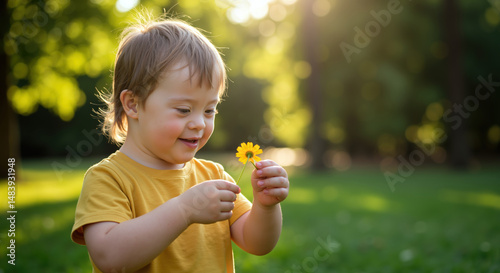 Boy with Down syndrome holding yellow flower in sunny park. Child in yellow t-shirt examining daisy with joy. Inclusion concept. Special needs childhood. Nature discovery. Horizontal banner