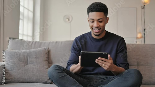 A joyful young man sits on a sofa in his apartment looking at a tablet. Represents happy home technology use