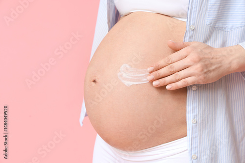 Pregnant woman applying body cream on her belly on pink background, closeup