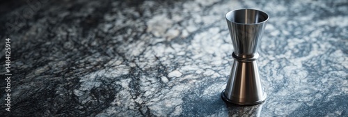 A sleek, stainless steel jigger rests on a dark, mottled countertop.  The minimalist setting accentuates the jigger's simple elegance.