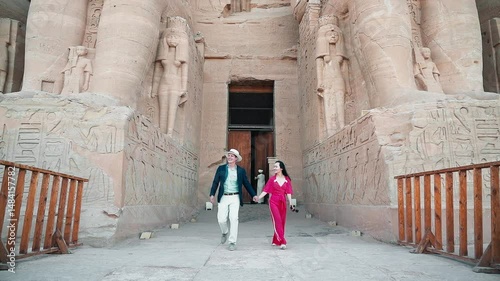 american young man traveler wearing white hat with his wife with red dress exploring ancient Egyptian abu simble temple with background of ramsses ii statues. Egypt.