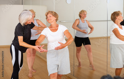 Elderly women who are engaged in the dance section at a group lesson stand in the 1st position of the ballet stand, where ..the choreographer helps them to do it correctly