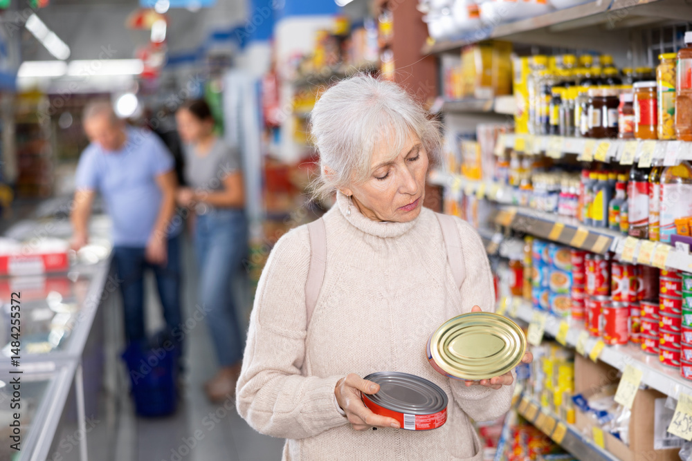 © JackF - Focused interested elderly woman standing in grocery store aisle carefully reading nutritional information on tin cans while choosing right canned food..