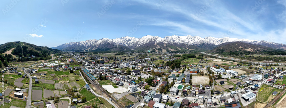 Naklejka premium Panoramic view of a peaceful rural town nestled in a valley with snow-capped mountains towering in the background under a clear blue sky at Hakuba Mountain Nagano, Japan.