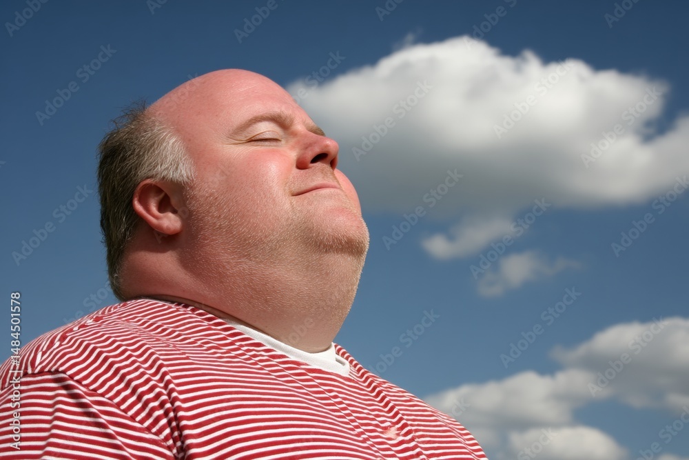 Obraz premium A man in a red striped shirt enjoys the sunshine outdoors, eyes closed, with a peaceful expression against a blue sky with clouds.