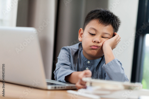 Bored Asian young boy sitting at desk with laptop, looking disinterested and resting his head on his hand. room has natural light and calm atmosphere