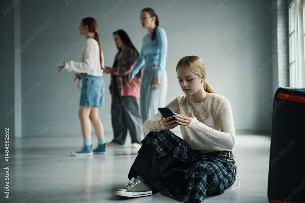 Fototapeta premium Teenager sitting on floor focused on smartphone with friends practicing dance in background in spacious room with natural light streaming through windows