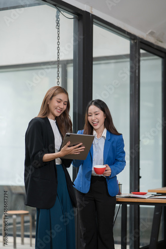 Two  Asian businesswomen holding iPads, holding graph papers, standing in front of the glass, talking and consulting with each other.