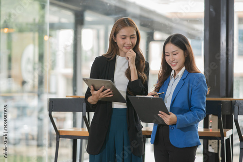 Two  Asian businesswomen holding iPads, holding graph papers, standing in front of the glass, talking and consulting with each other.