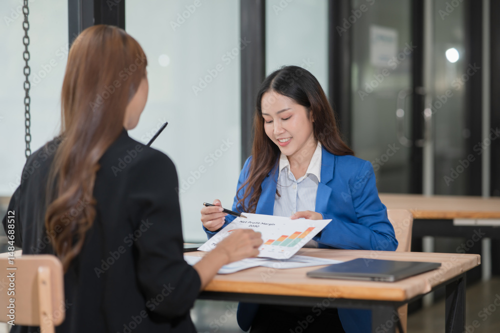 Two Asian businesswomen discuss chart documents in an office.