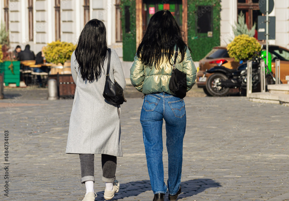 Naklejka premium Rear view of two stylish and elegant brunette young women walking on the street in the town square on a sunny spring day. Two attractive female friends walking in the downtown. Urban street life scene