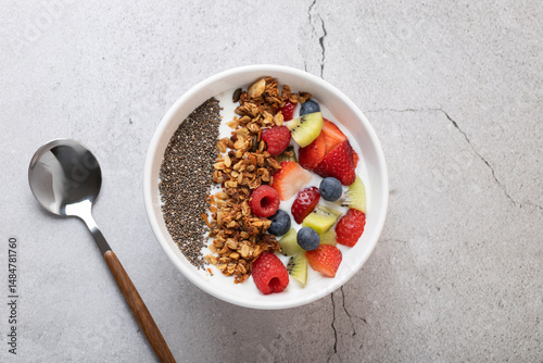 Top view of white bowl with kefir, strawberries, kiwi, raspberries, blueberries, chia seeds and granola, with metal and wood spoon on gray stone background. 