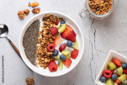 Top view of white bowl with kefir, granola, chia and fresh fruits. On the left, a metal spoon; on the right, an open granola jar and a bowl of fruit. Concept of natural breakfast and unprocessed food