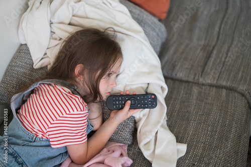 Cute sad girl in denim overalls lying on the sofa with remote control. Concept of children's emotions and leisure