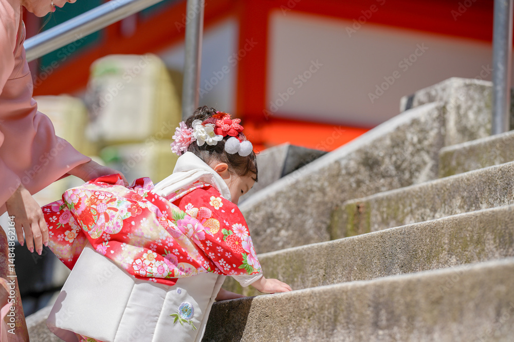 Obraz premium A 3-year-old Japanese girl in a red kimono climbs shrine steps during Shichi-Go-San, helped by her mom in a pink kimono. This photo captures a tender traditional moment between mother and daughter.