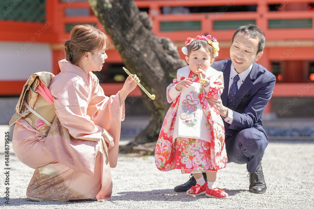 A 3-year-old Japanese girl in a red kimono plays under cherry blossoms at a shrine with her parents-mom in a pink kimono, dad in a suit-during Shichi-Go-San. This photo shows a joyful family moment. A 3-year-old Japanese girl in a red kimono plays under cherry blossoms at a shrine with her parents-mom in a pink kimono, dad in a suit-during Shichi-Go-San. This photo shows a joyful family moment.
