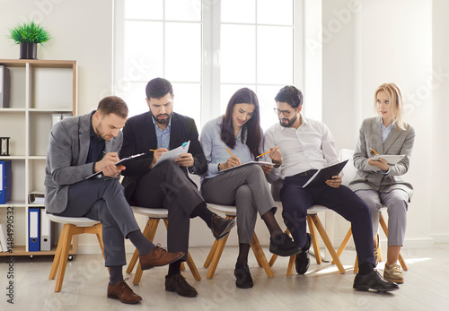 Group of young people sitting in row on chairs, holding paper documents and preparing for job interview or having training business session, waiting in queue, discussing some things