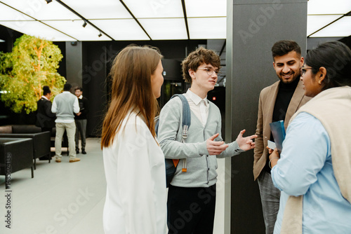 A multinational group of young people in business attire is standing indoors in a modern office lobby during an internship program, with a young White man holding notebooks and gesturing.