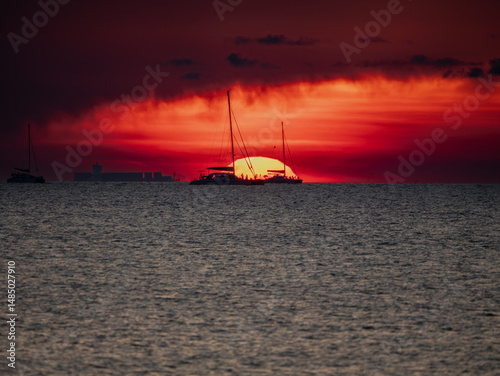 Silhouettes of sailboats and a cargo ship during fiery sunset at Sunset Village Beach Resort, Najomtien, Chon Buri, Thailand.