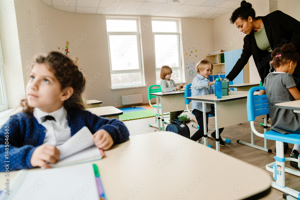 Fototapeta premium An adult Black female teacher assisting a multinational group of young pupils sitting at their desks in a bright modern classroom with large windows, blue chairs, and colorful educational decorations.