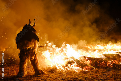 A Krampus figure stands before a large bonfire, smoke fills the air. Krampus and St Nicholas, 5 December in Tarvisio, Friuli Venezia Giulia, Julian Alps, Italy.