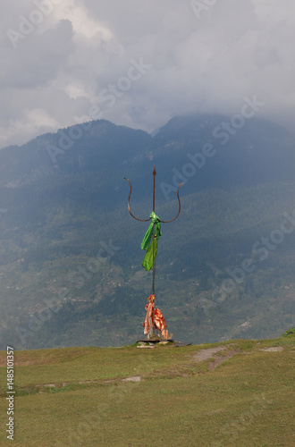 Majestic Shiva's trident adorned with holy cloth stands tall against a misty mountain backdrop and green meadow