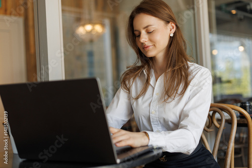 Young professional woman in a white shirt working on a laptop at a modern cafe. Confident and smiling, she represents success, remote work, and a stylish business lifestyle. Free Download Young professional woman in a white shirt working on a laptop at a modern cafe. Confident and smiling, she represents success, remote work, and a stylish business lifestyle. - GFXLoad