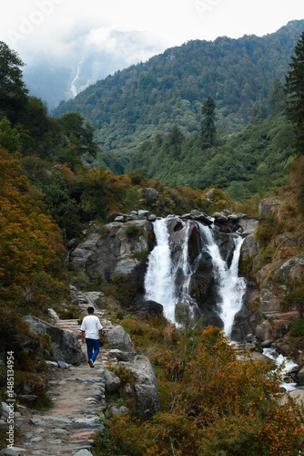 A lone traveler walks along a scenic trail toward a cascading waterfall nestled in lush, misty mountains.