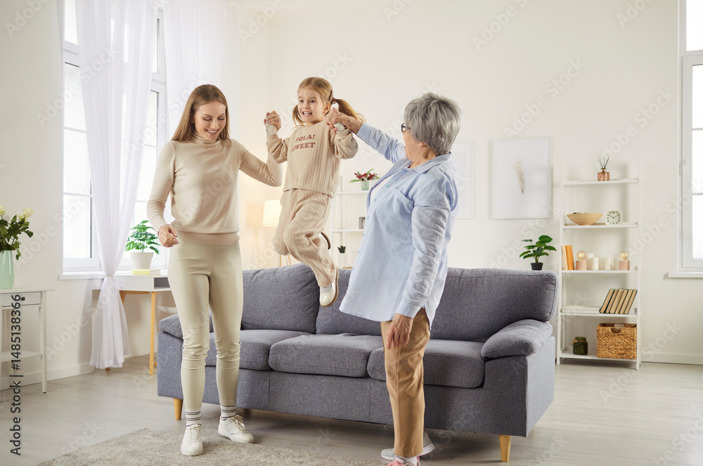 Obraz premium Three female generations have fun together, spend spare time at home. Little girl jumps and holds hands of her mother and grandmother, plays actively. Family and relationship concept