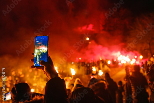 A hand holds a smartphone, recording a crowd with torches against a red sky. The phone screen shows the same scene. Krampus and St Nicholas, 5 December in Camporosso Valcanale, Tarvisio, Italy.