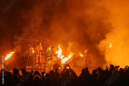 Crowd watches figures in fur costumes and horned masks holding torches amidst smoke and fire. Krampus and St Nicholas, 5 December in Camporosso Valcanale, Tarvisio, Italy.