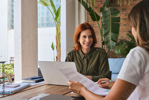 Happy busy young smiling professional business woman employee, financial advisor or company manager consulting female client, talking to colleague working in office using laptop sitting at desk.