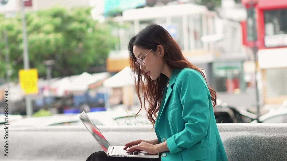 A woman in a teal blazer works on a laptop outdoors in an urban area during the day. A woman in a teal blazer works on a laptop outdoors in an urban area during the day.
