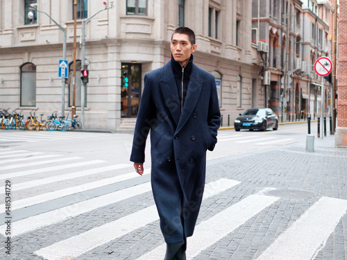 Portrait of handsome Chinese young man wearing black overcoat posing in the street, young guy with black short hair all in black with urban background. Male fashion, cool Asian young man lifestyle.