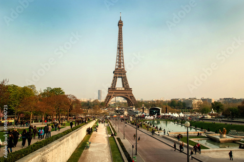 Horizontal view of the Eiffel Tower in Paris, France, captured head-on with the lush lawns of the Champ de Mars in the foreground. The iconic iron structure stands tall under a clear blue sky, framed 