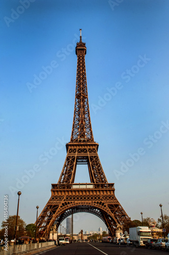 Horizontal view of the Eiffel Tower in Paris, France, captured head-on with the lush lawns of the Champ de Mars in the foreground. The iconic iron structure stands tall under a clear blue sky, framed 