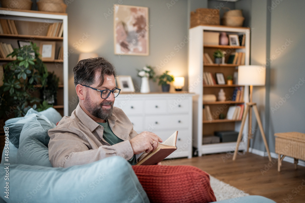 Fototapeta premium Smiling bearded man reading a book at home on the sofa