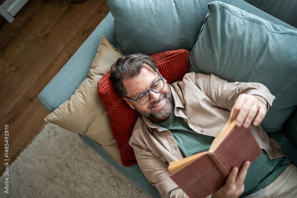 Fototapeta premium Smiling man lying on sofa reading a book at home
