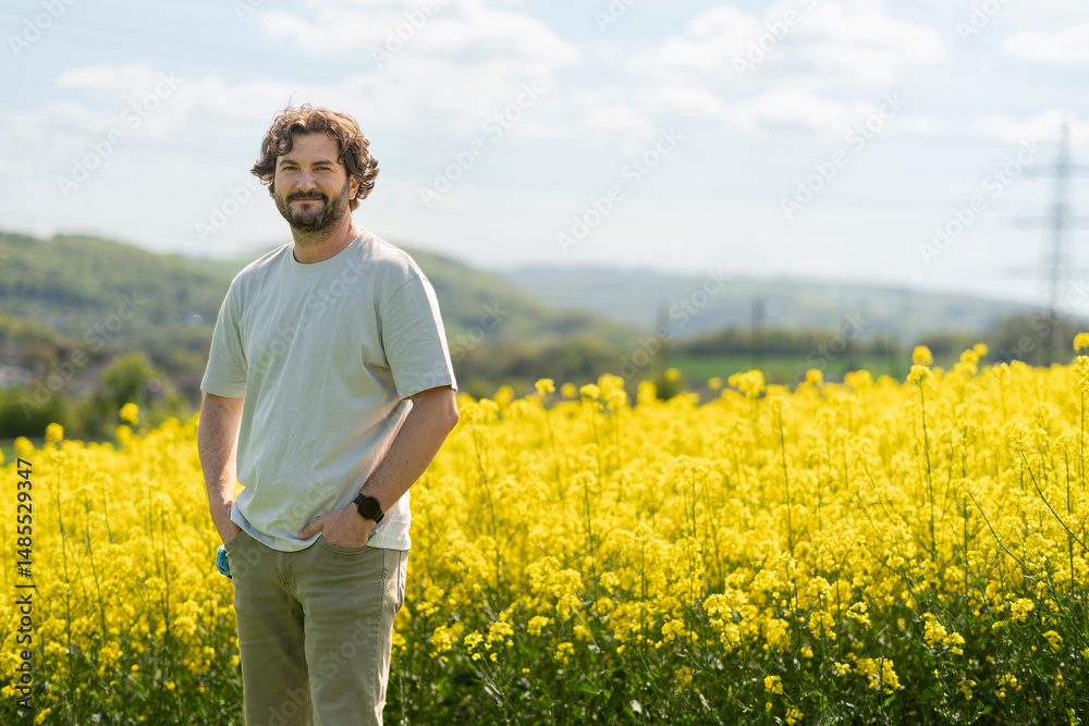 Fototapeta premium Cute bearded curly man in T-shirt smiling standing in front of yellow blooming rapeseed field. Concept of emotions, leisure and travel