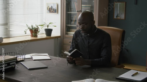African American catholic priest wearing glasses and black clergy shirt with white collar reading small bible book and holding rosary while sitting at desk in office