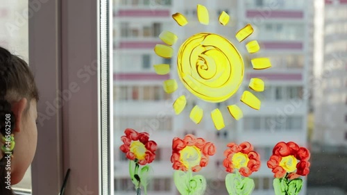 Little girl with brush in hand painting flowers and sun on the window at home. Colorful education at home, fun and play sensory 